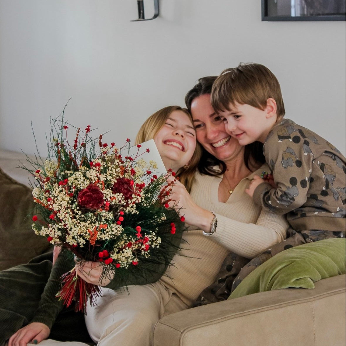 En glad mamma med sina barn i soffan, håller en Mors dag-bukett och läser ett kort.