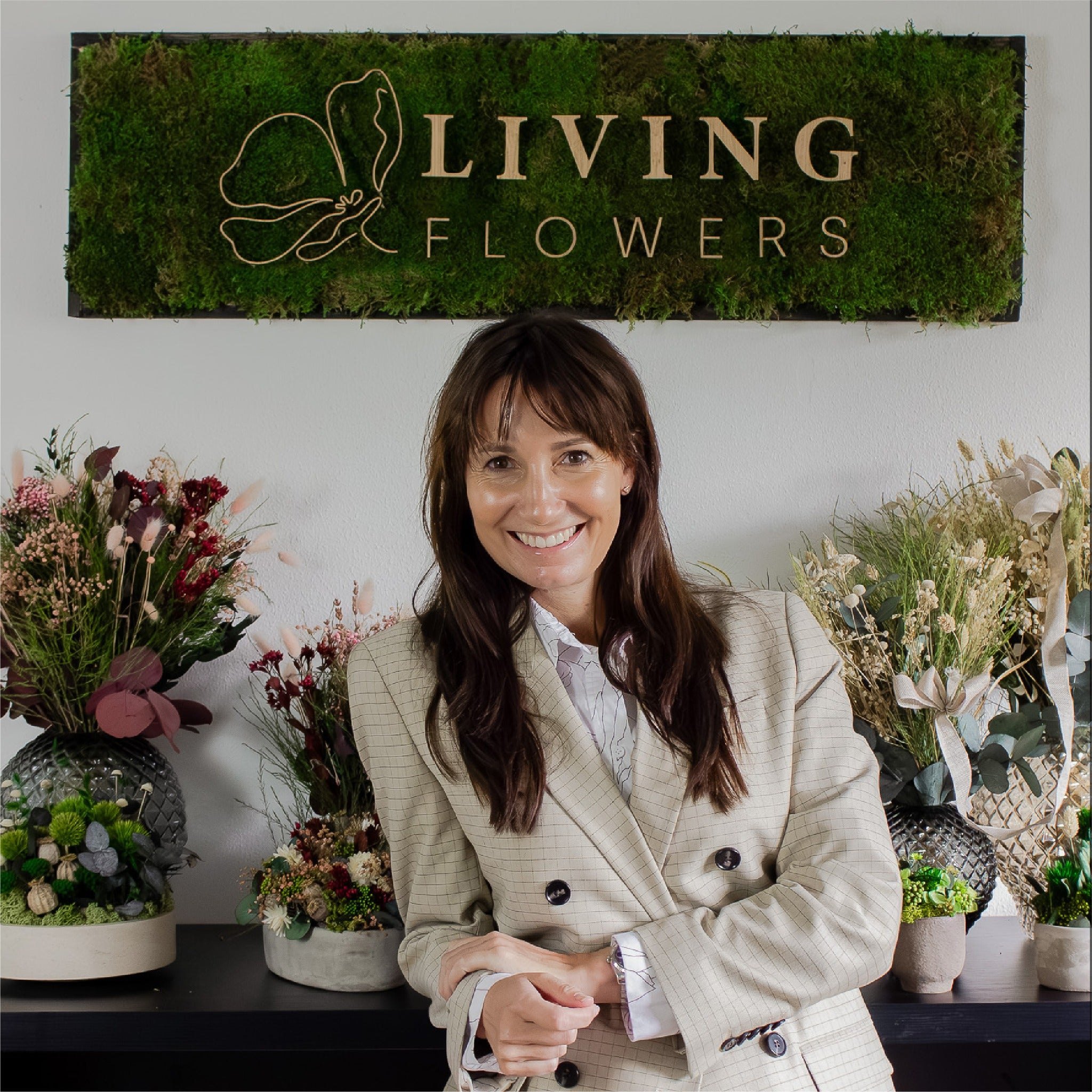 Britta - en av Living Flowers grundare står lutad mot ett högt bord fyllt av olika blomsterkreationer.