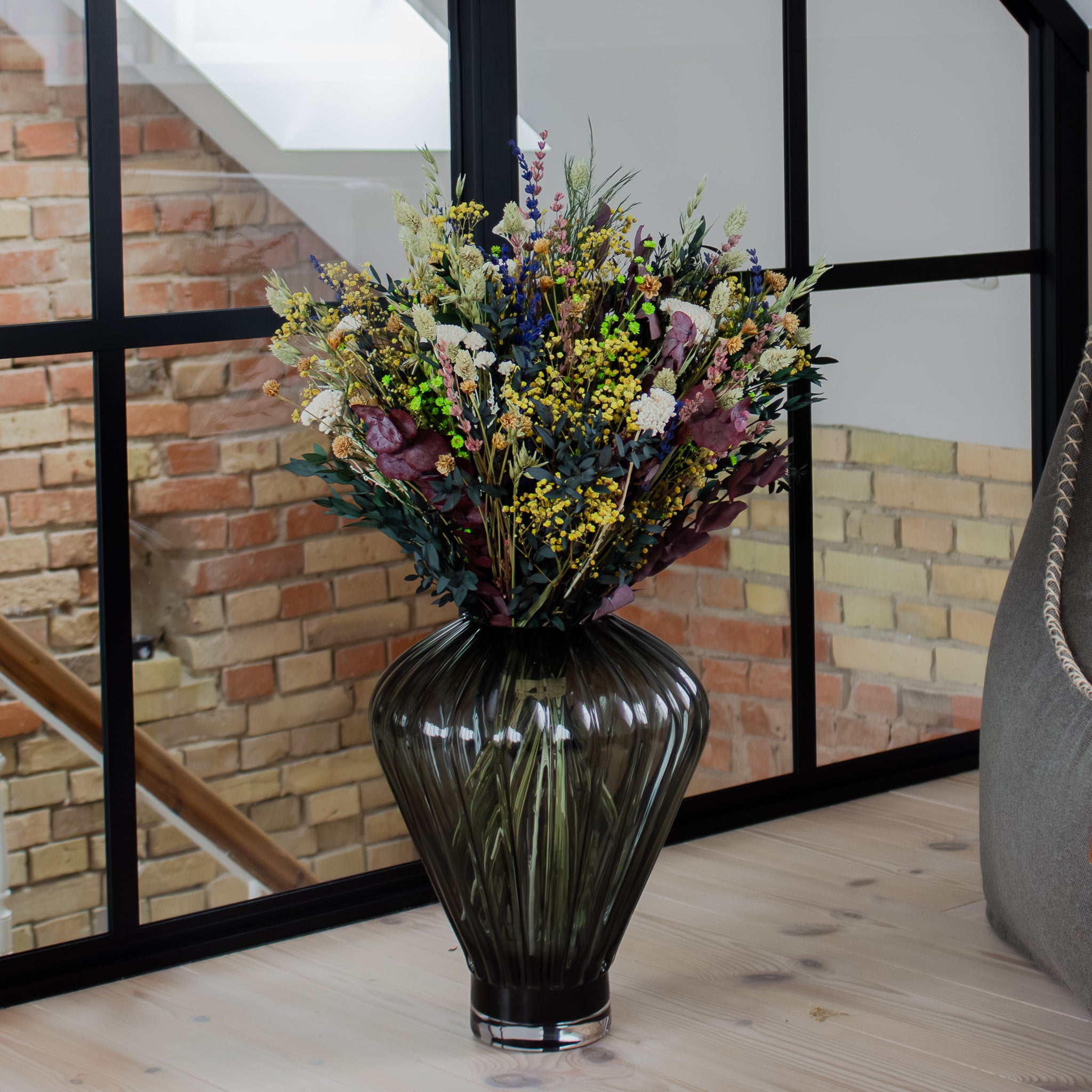 Blombukett i gult, beige, blått, vitt och rosa står i en vacker glasvas på ett trägolv.