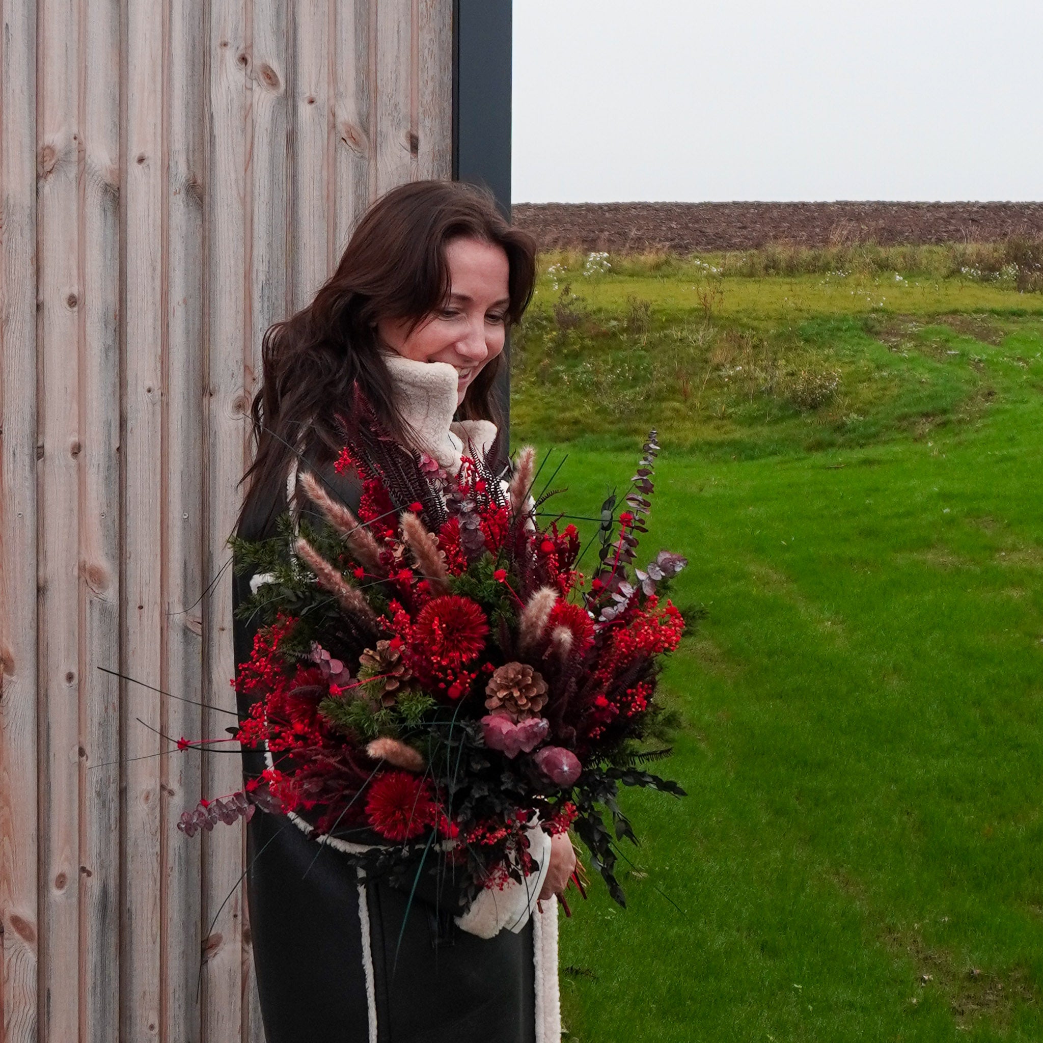 Utomhusbild på kvinna som håller en mörkröd bukett vid en husvägg i trä.