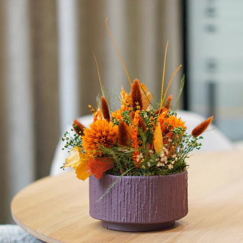 Närbild av en orange blomdekoration i en lila skål, som står på ett träbord.