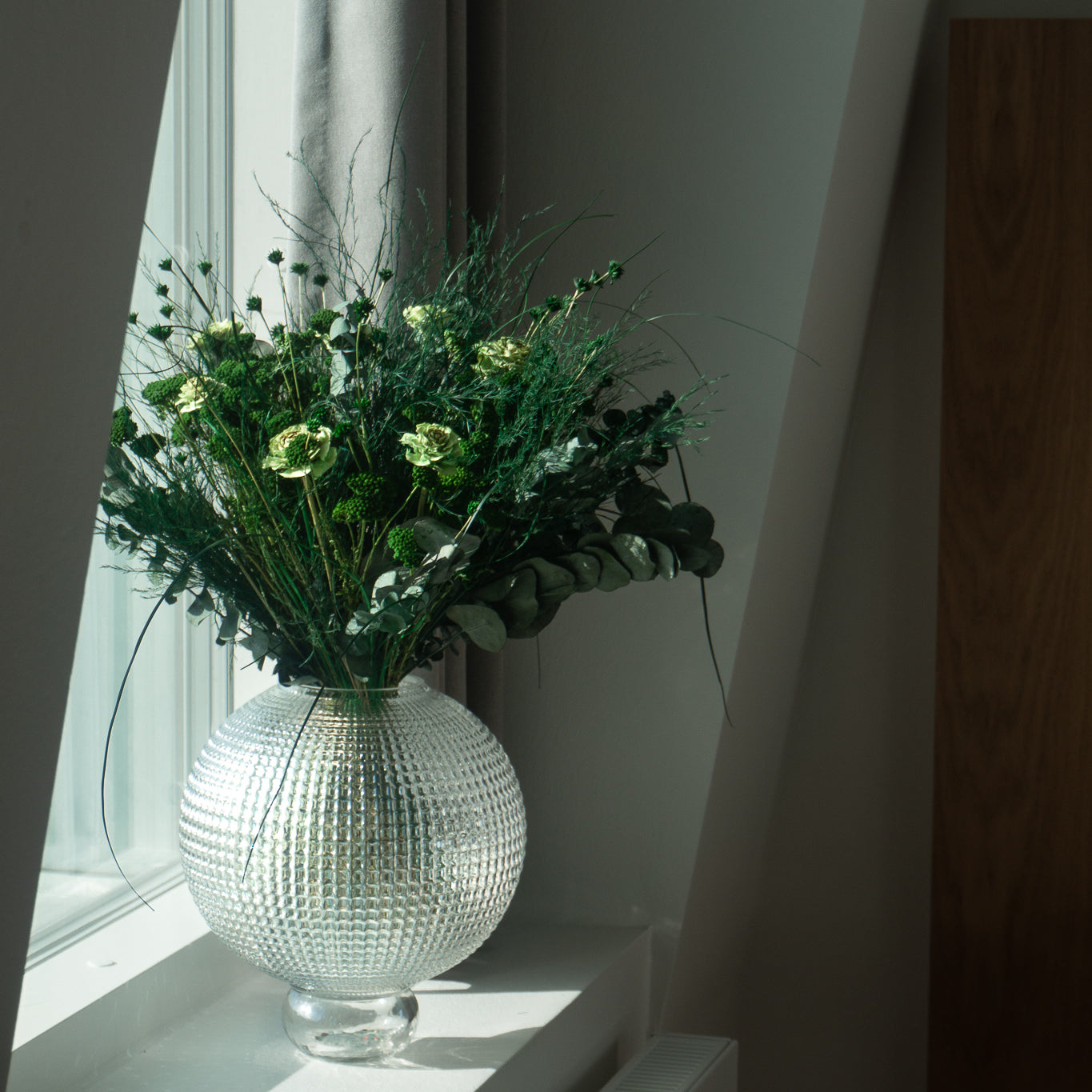 Grön bukett i en stor, rund glasvas placerad på en fönsterbräda.