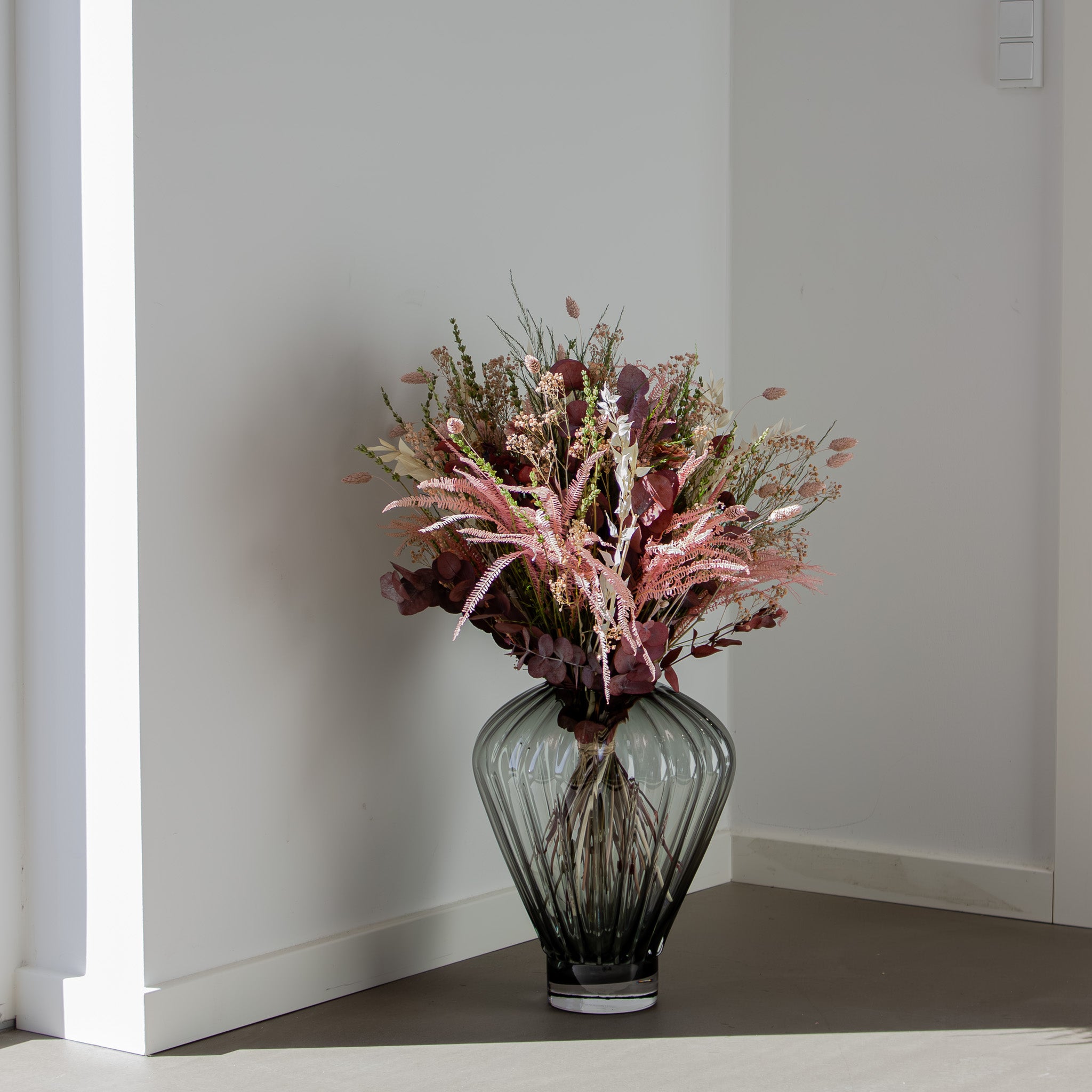 En vacker blombukett i rosa toner som står placerad i en grå glasvas, på ett mörkt golv.