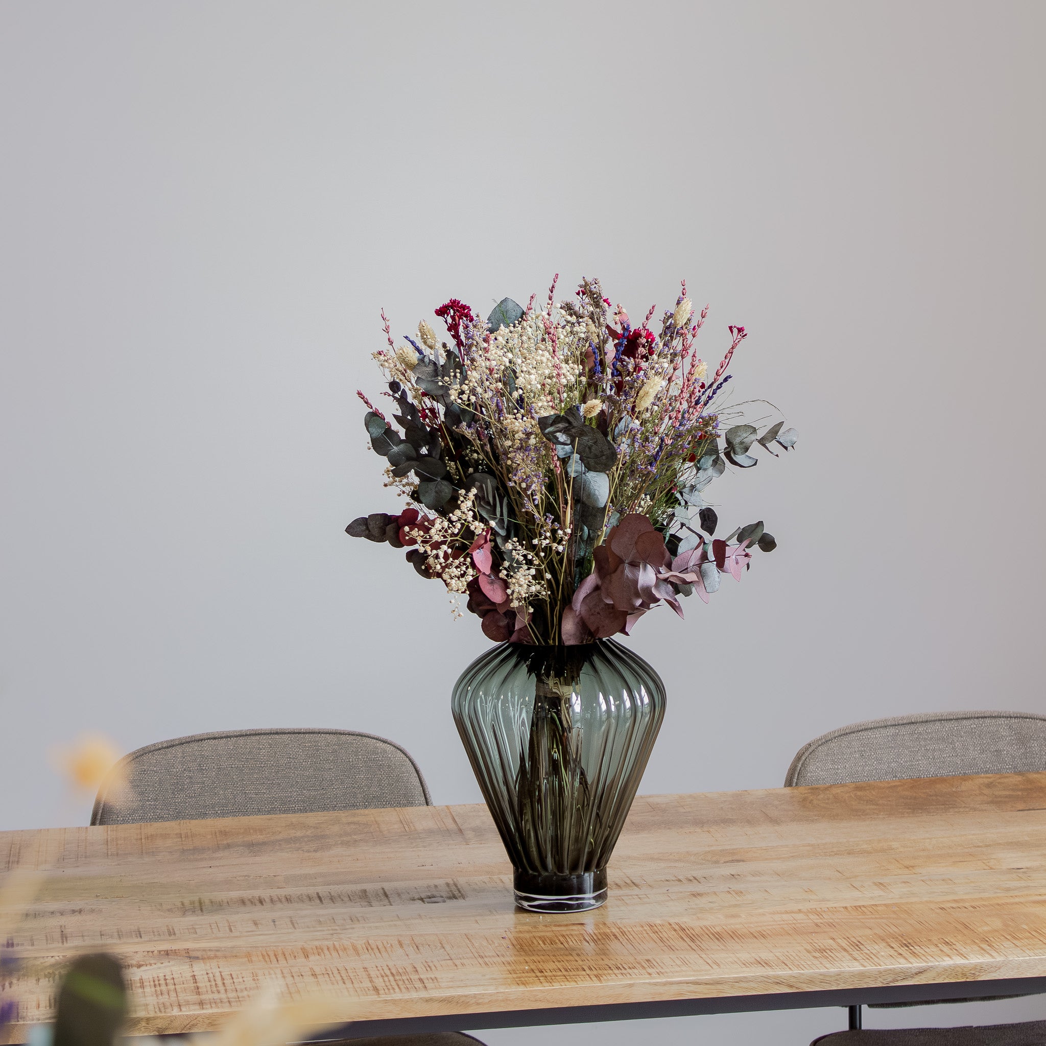 Bukett Frisk Fläkt i dova nyanser av rosa, grön, vit och blå placerad i en glasvas på ett stort träbord