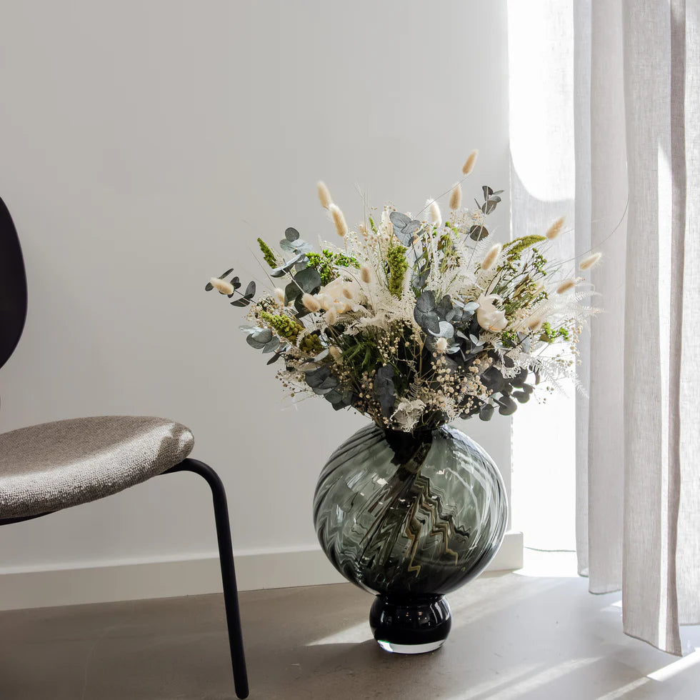 Buketten Grön Harmoni i grönt och vitt står i en vacker rund glasvas på ett betonggolv, med en beige gardin bakom.