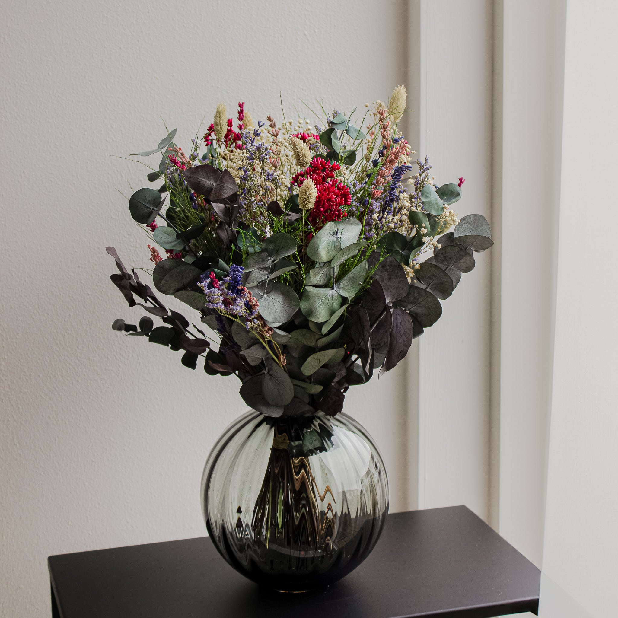 Bukett Frisk Fläkt i dova färger av grönt, lila, rosa och beige- i en vacker glasvas på ett litet bord