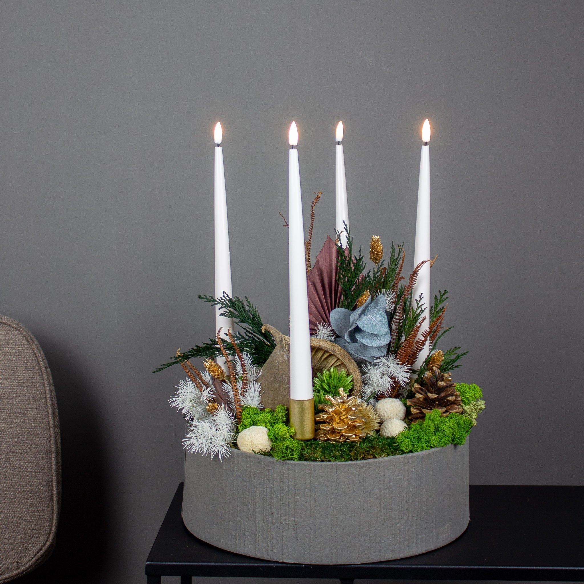 Et julearrangement med mos, kogler, langtidsholdbare blomster og fire lys er arrangeret i en skål.