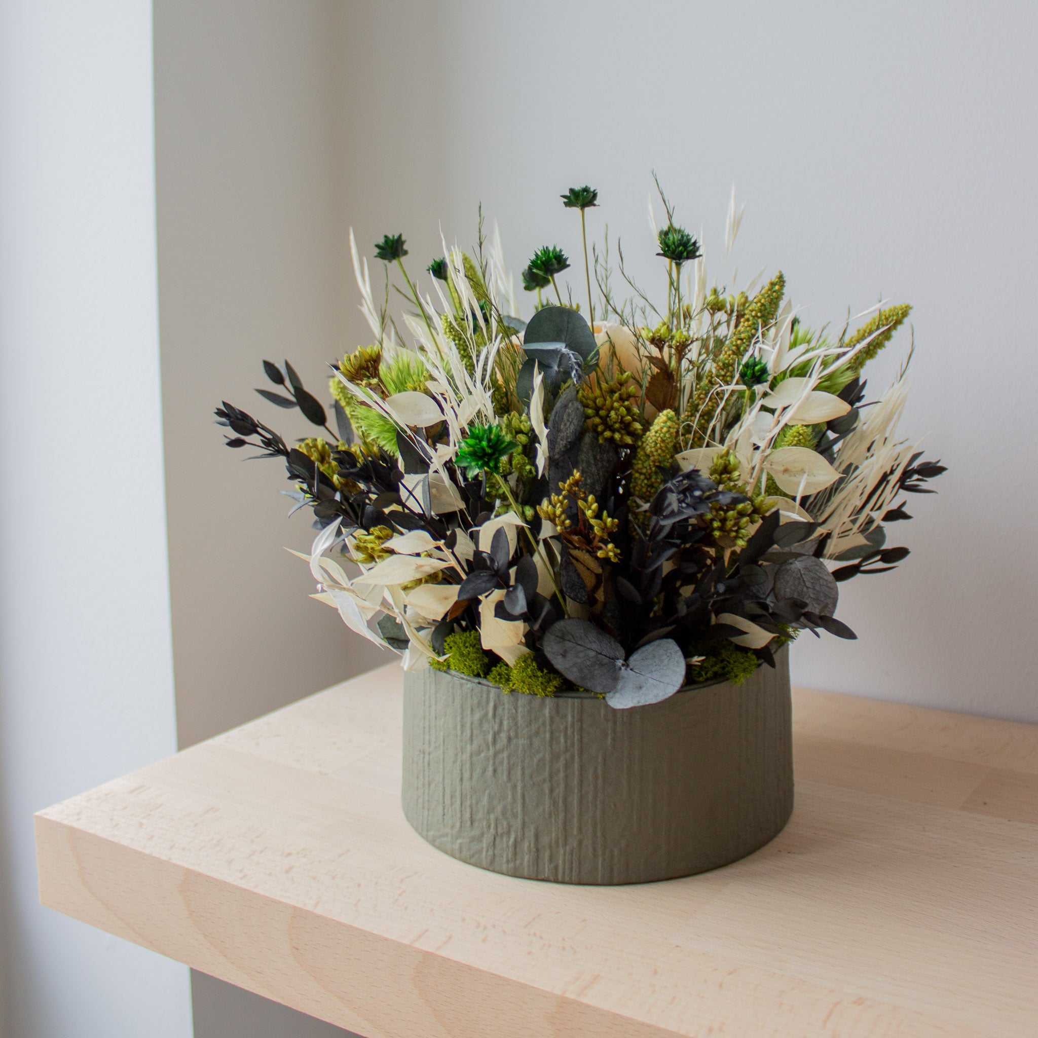 Stilren blomdekoration i grönt och vitt, placerad i en grå papp-kruka på en träbänk