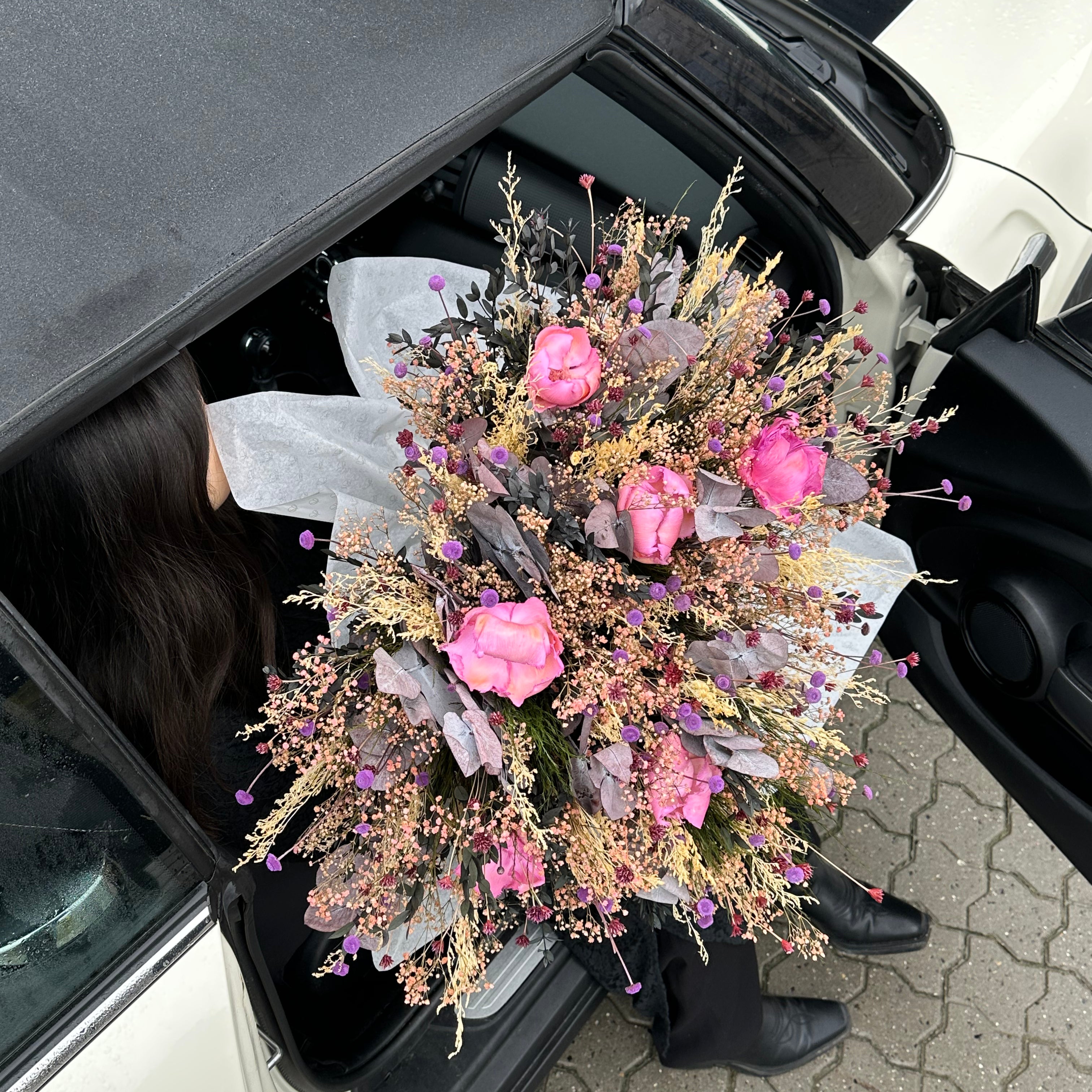 Kvinna som är påväg ut ur sin bil med en stor blomsterbukett i armarna - i rosa och lila nyanser.