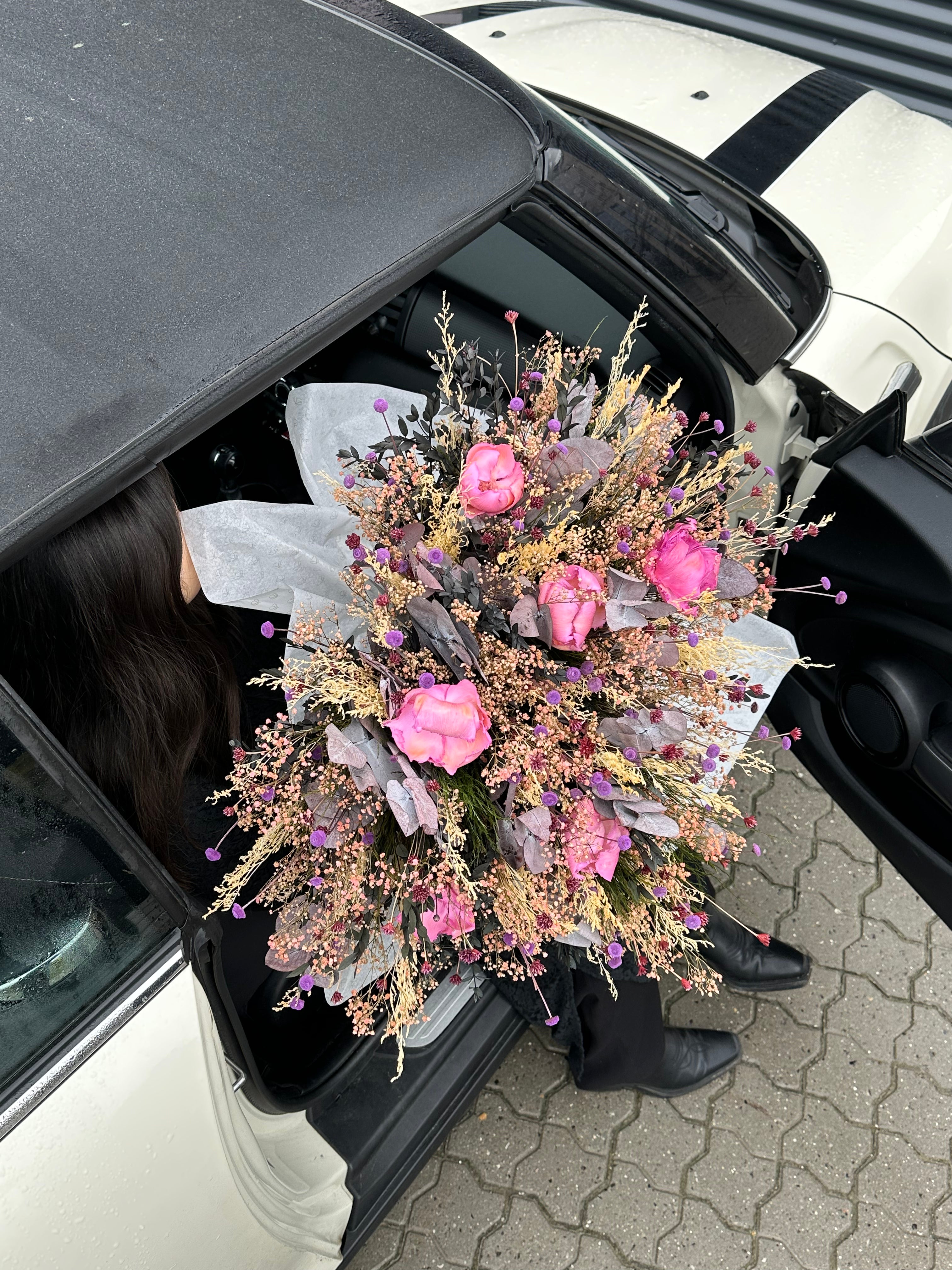 Kvinna som är påväg ut ur sin bil med en stor blomsterbukett i armarna - i rosa och lila nyanser.