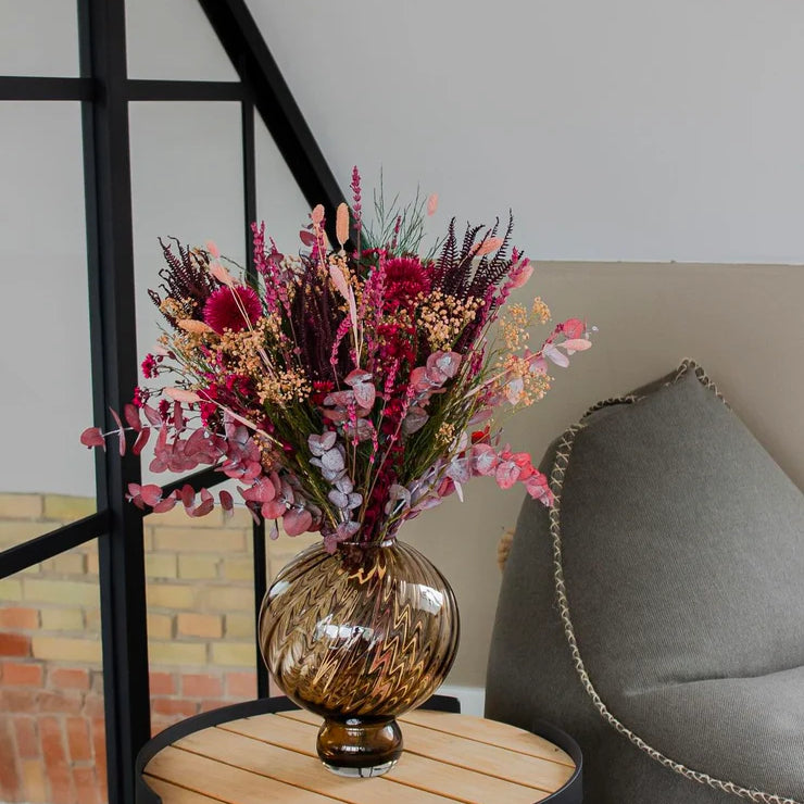 Blombukett i röda och rosa toner placerad i en brun glasvas på ett litet runt bord