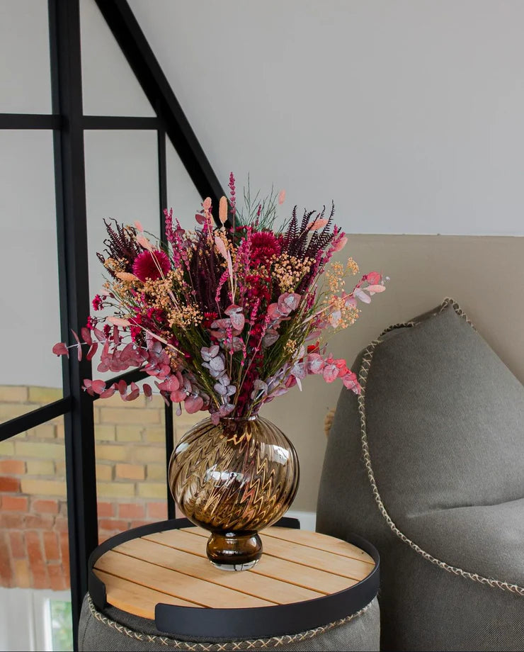 Blombukett i röda och rosa toner placerad i en brun glasvas på ett litet runt bord