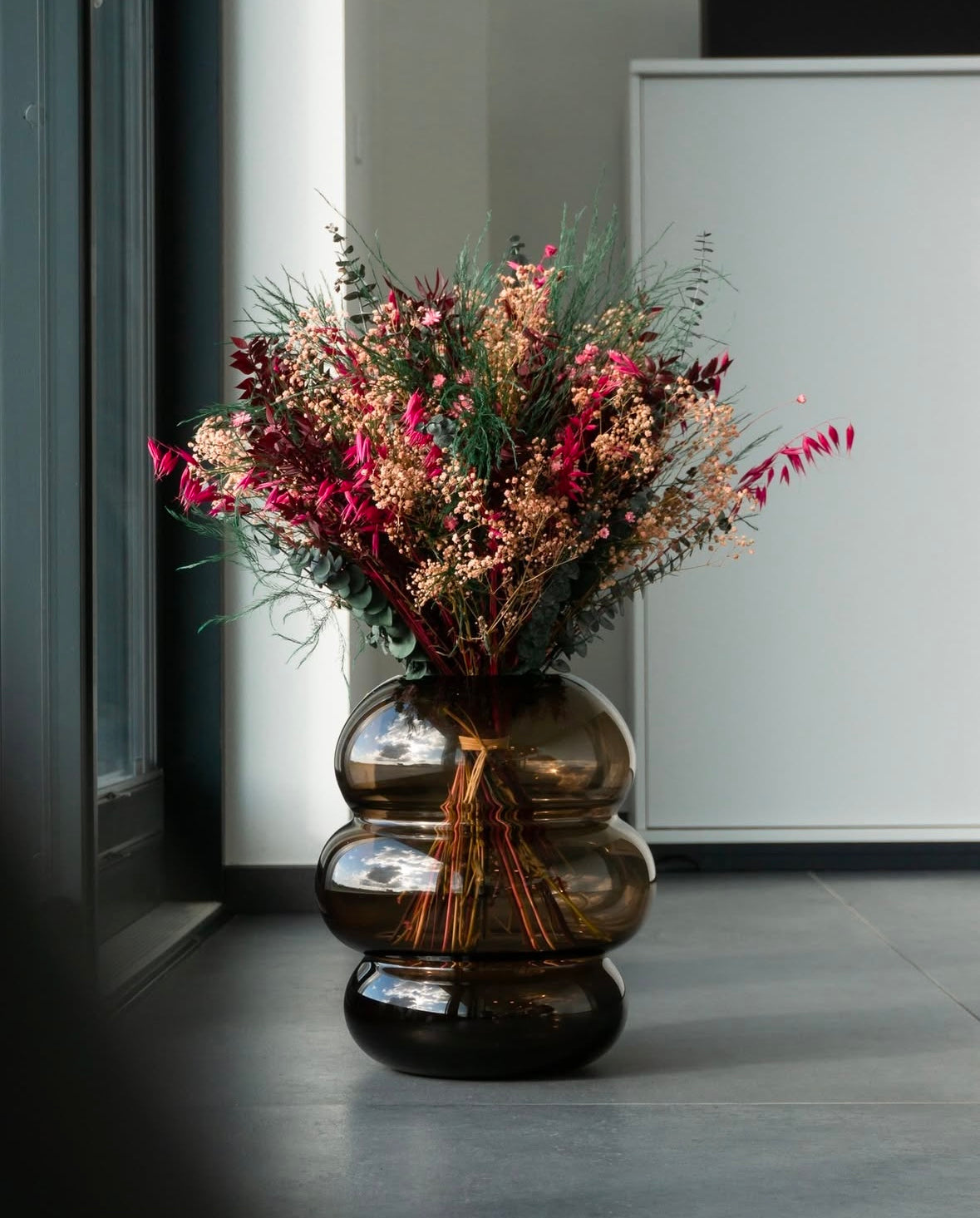 Brun, rosa och grön bukett placerad i stor brun glasvas på golvet.