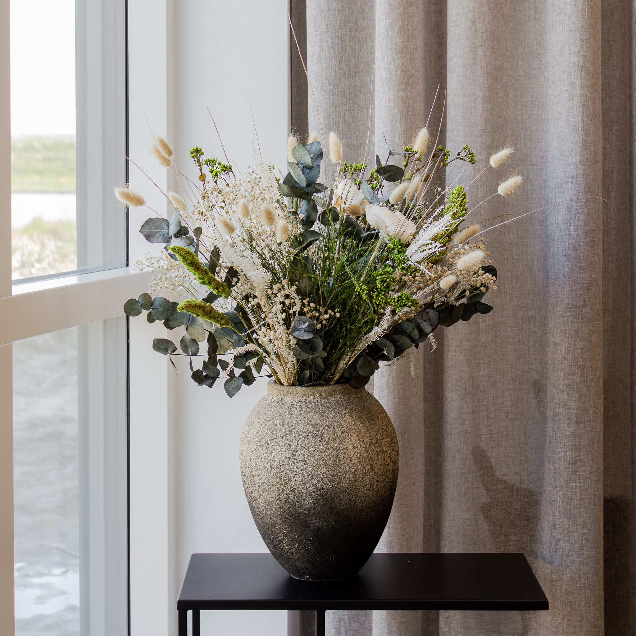 En grönvit blombukett i en fin, oval keramikvas framför ett fönster med gardin