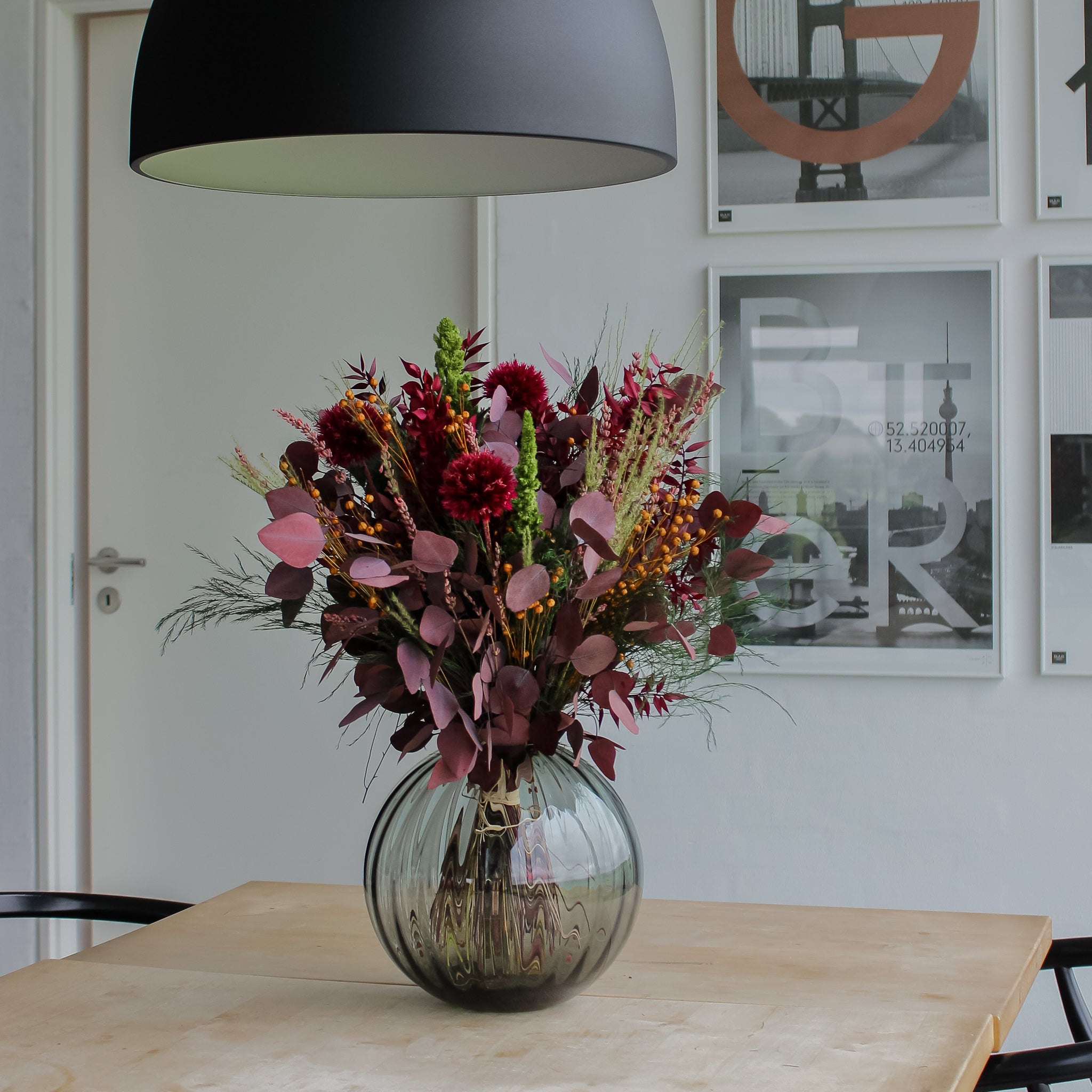 Blombukett i röda och gröna nyanser i glasvas placerad på ett träbord i hemmiljö.