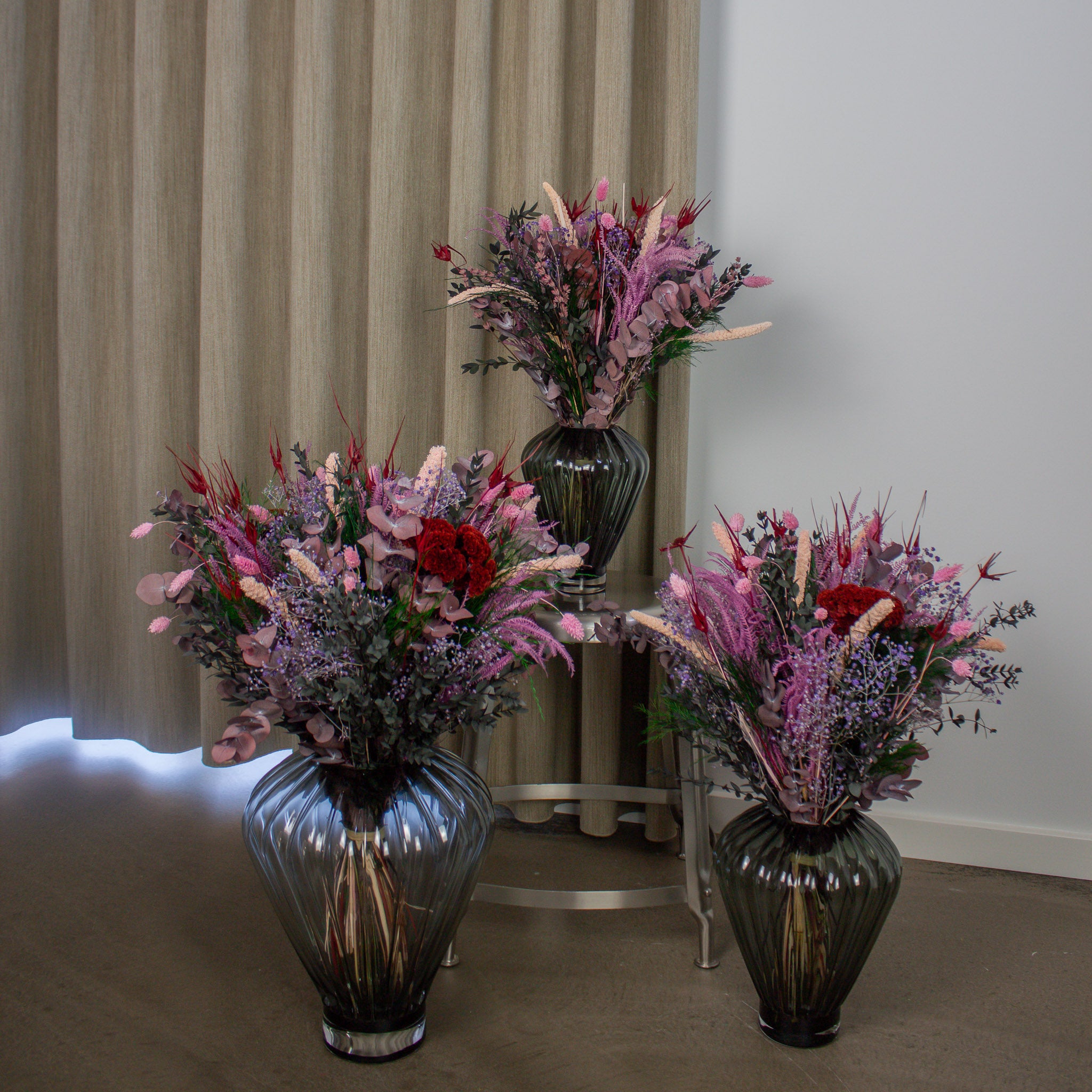 Vacker lila, röd och grön bukett i tre olika storlekar, där två av dem står på golvet medan den mittersta är placerad på en metallpall.