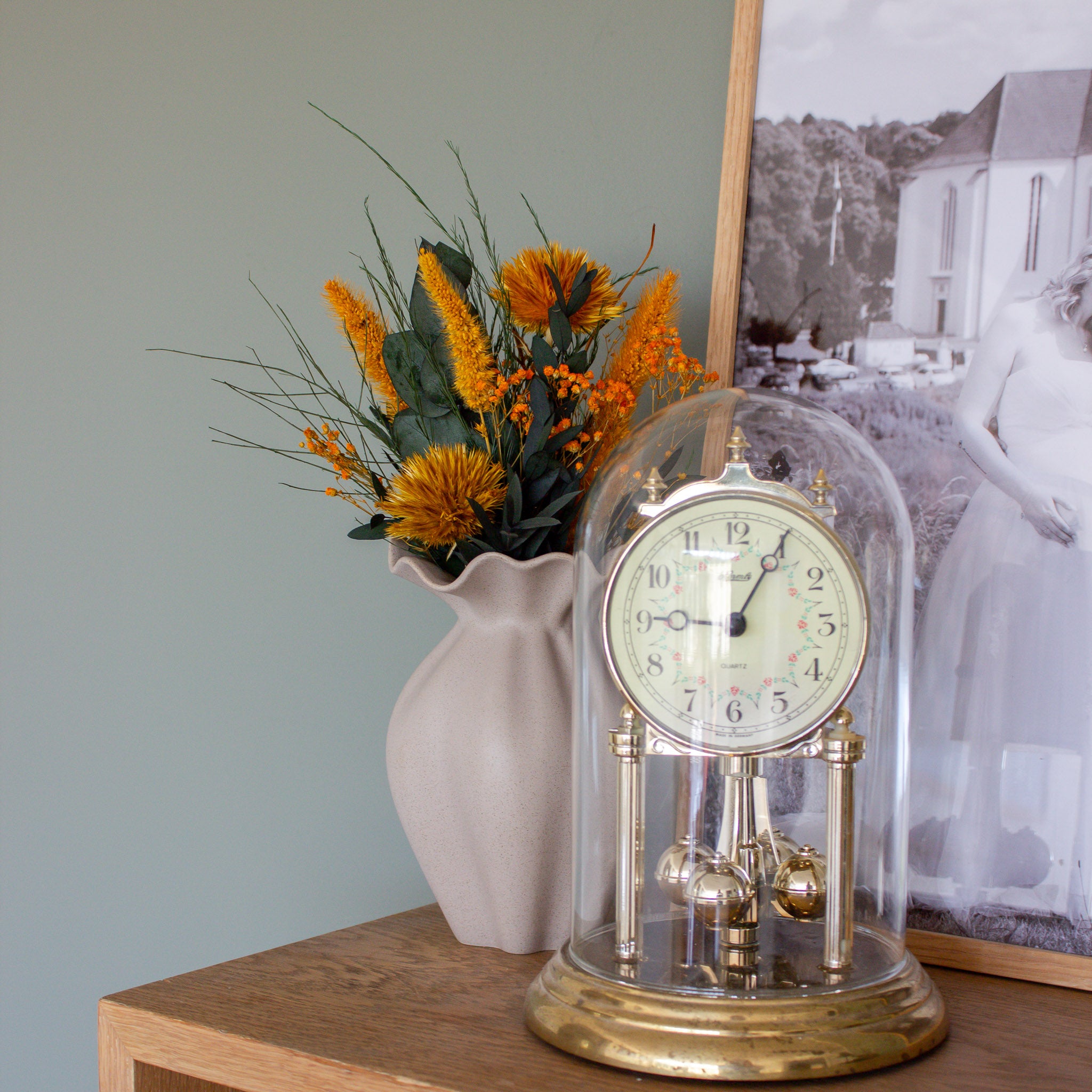 Stilleben med en gammal klocka, en tavla och Nellie-vasen med en orange miniatyrbukett.