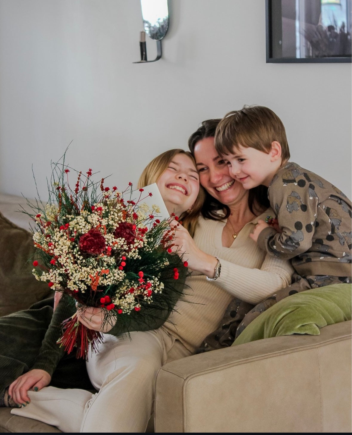 En glad mamma med sina barn i soffan, håller en Mors dag-bukett och läser ett kort.
