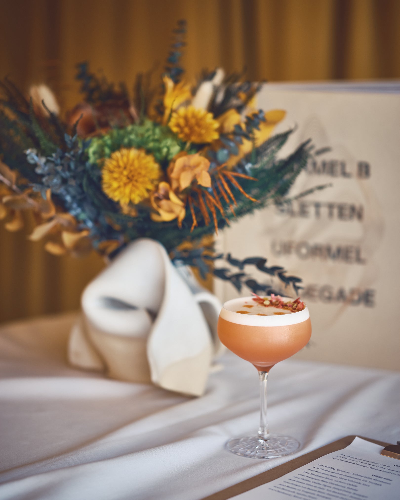 På et dækket bord med et fyldt cocktailglas står en efterårsbuket i varme orange-gule nuancer.