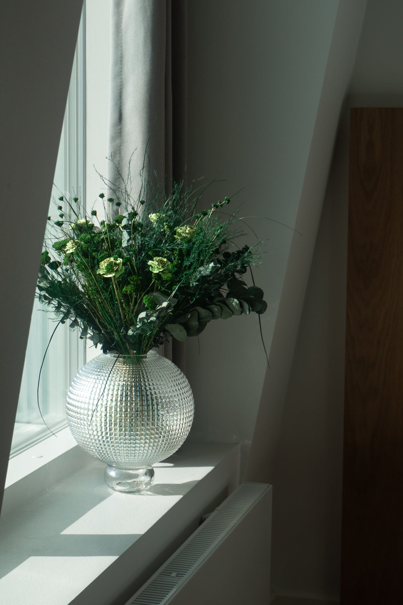 Bukket med langtidsholdbare blomster i stærke grønne nuancer står i en glasvase på en vindueskarm.