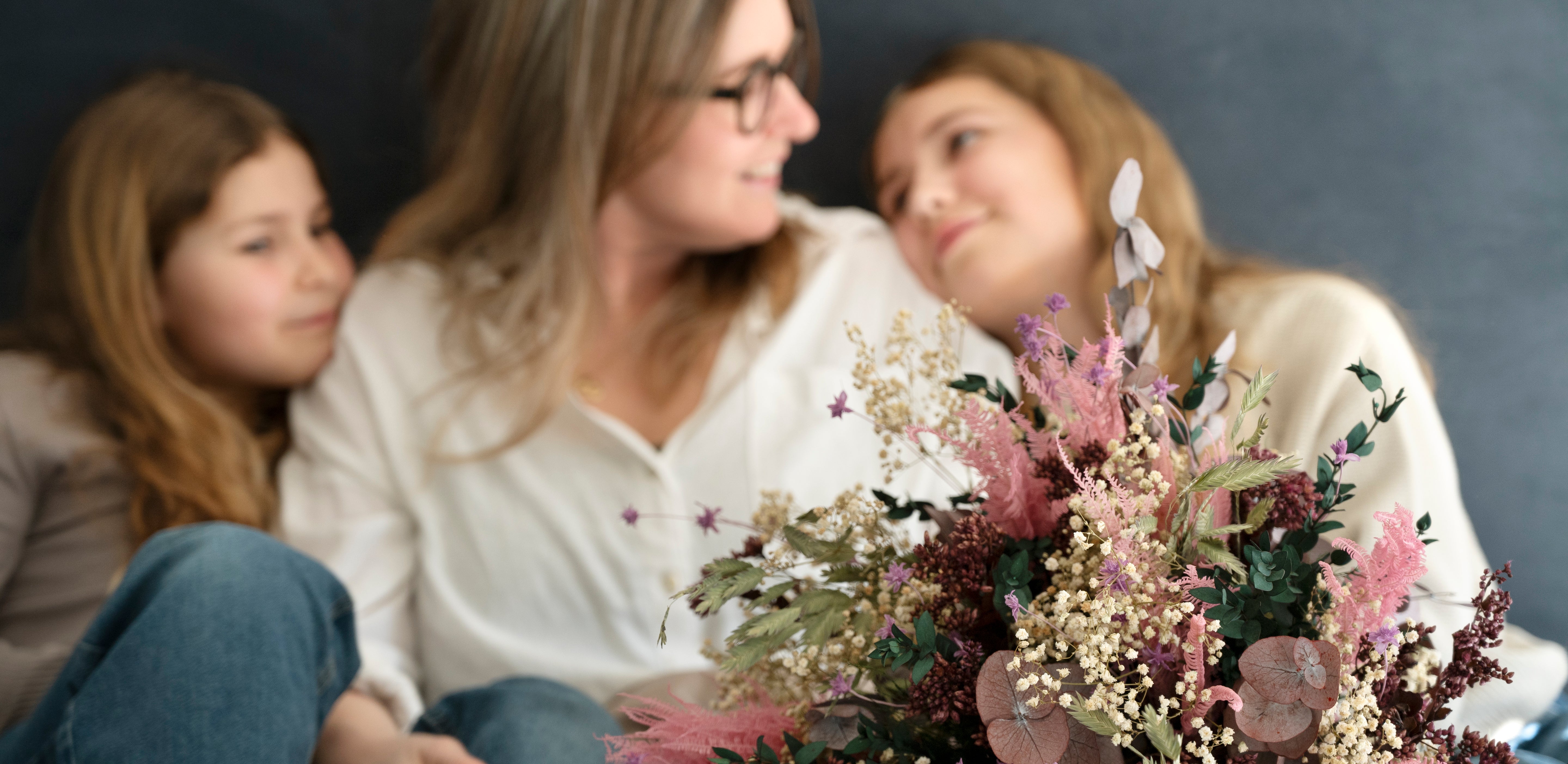 Fokus på en blombukett som ligger nära kameran och i bakgrunden syns en mamma som ler mot sina två barn