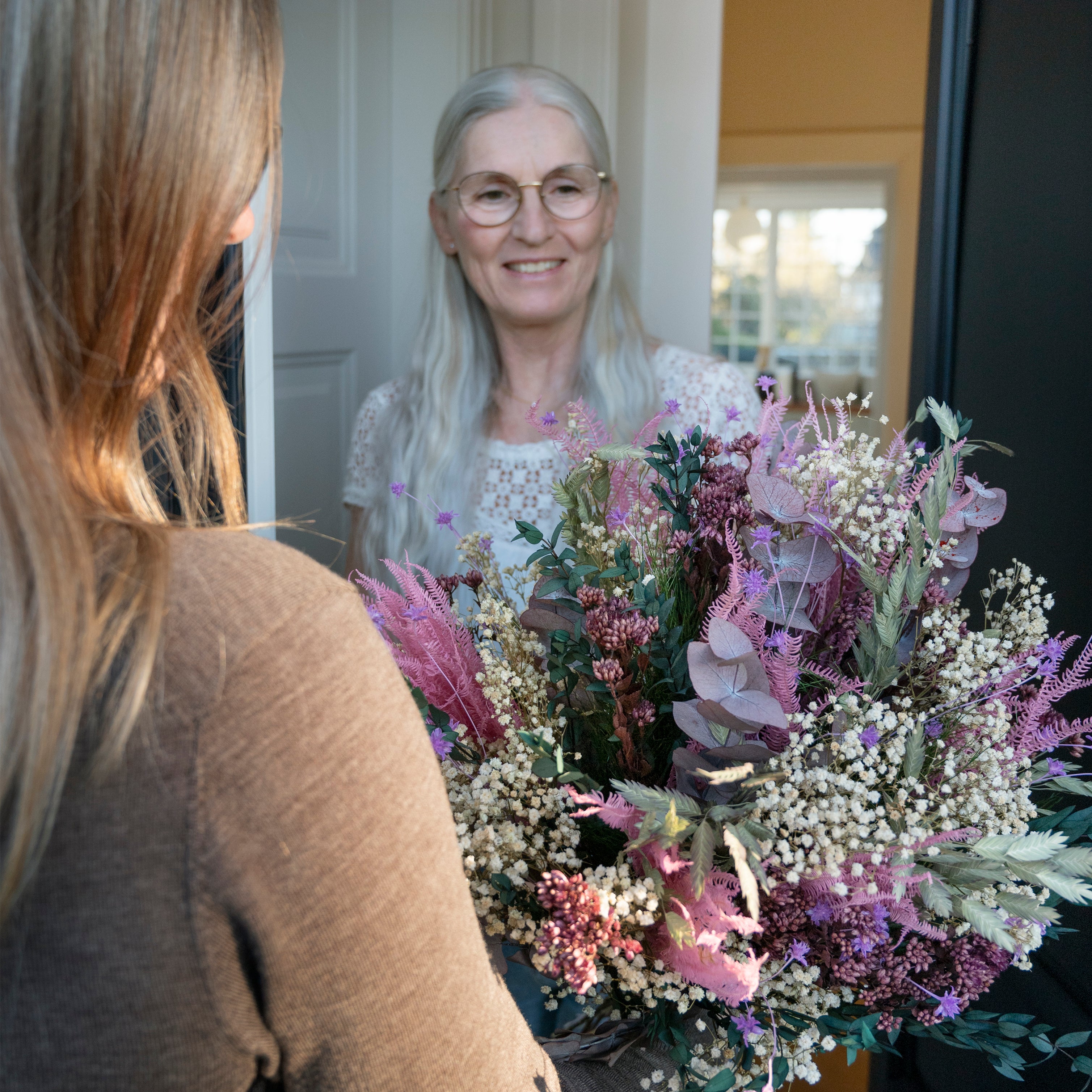 Mamma står i dörröppningen och ler när dottern räcker över en blombukett