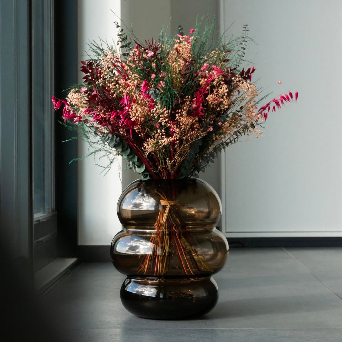 Stor buket i dybe grønne, rosa og rødviolette nuancer – arrangeret i en røgfarvet glasvase.