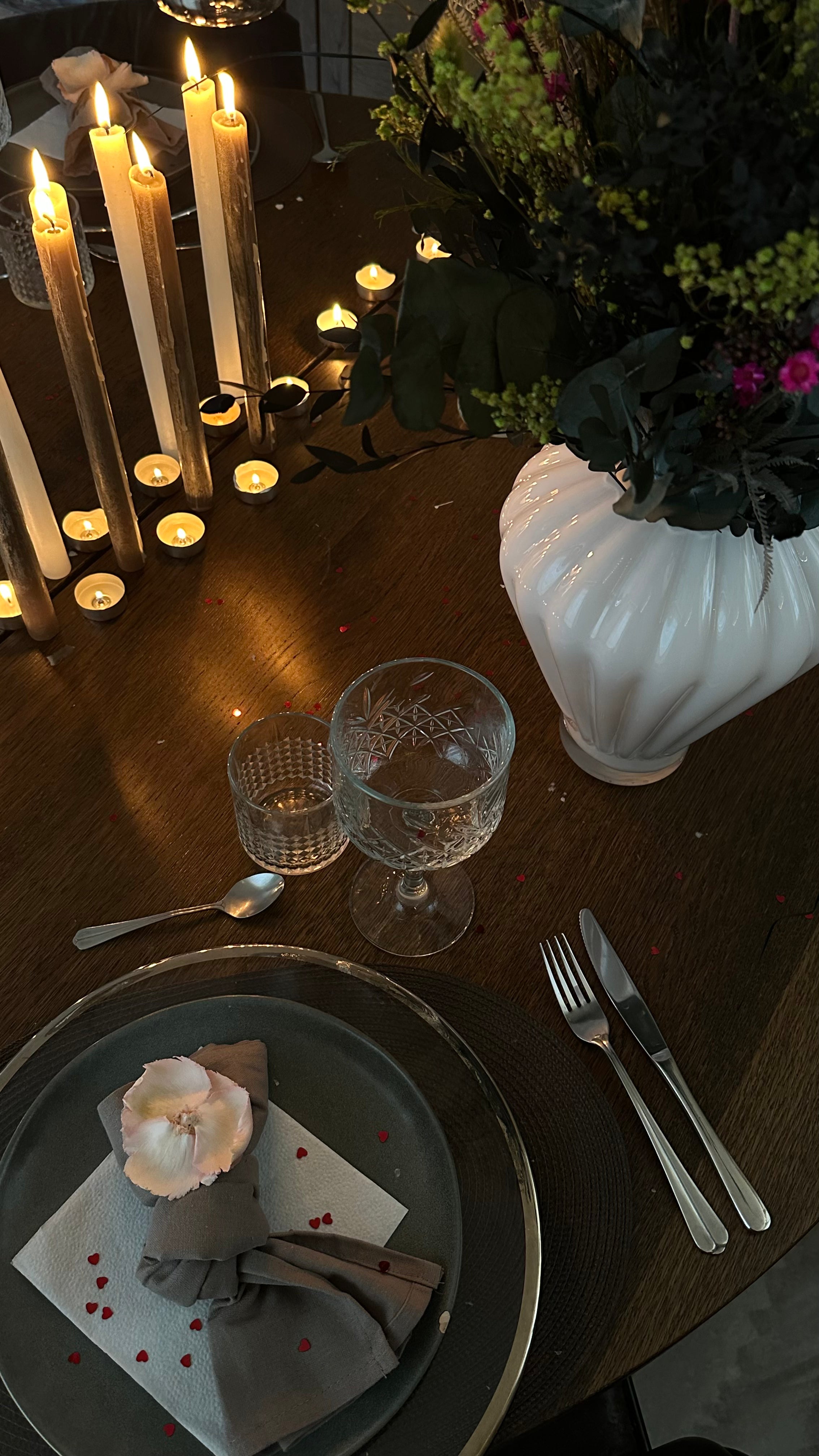Et smukt dækket bord i mørke nuancer med en stor buket i en elegant hvid vase. Stemningen forstærkes af mange levende lys og små røde hjertekonfettier.