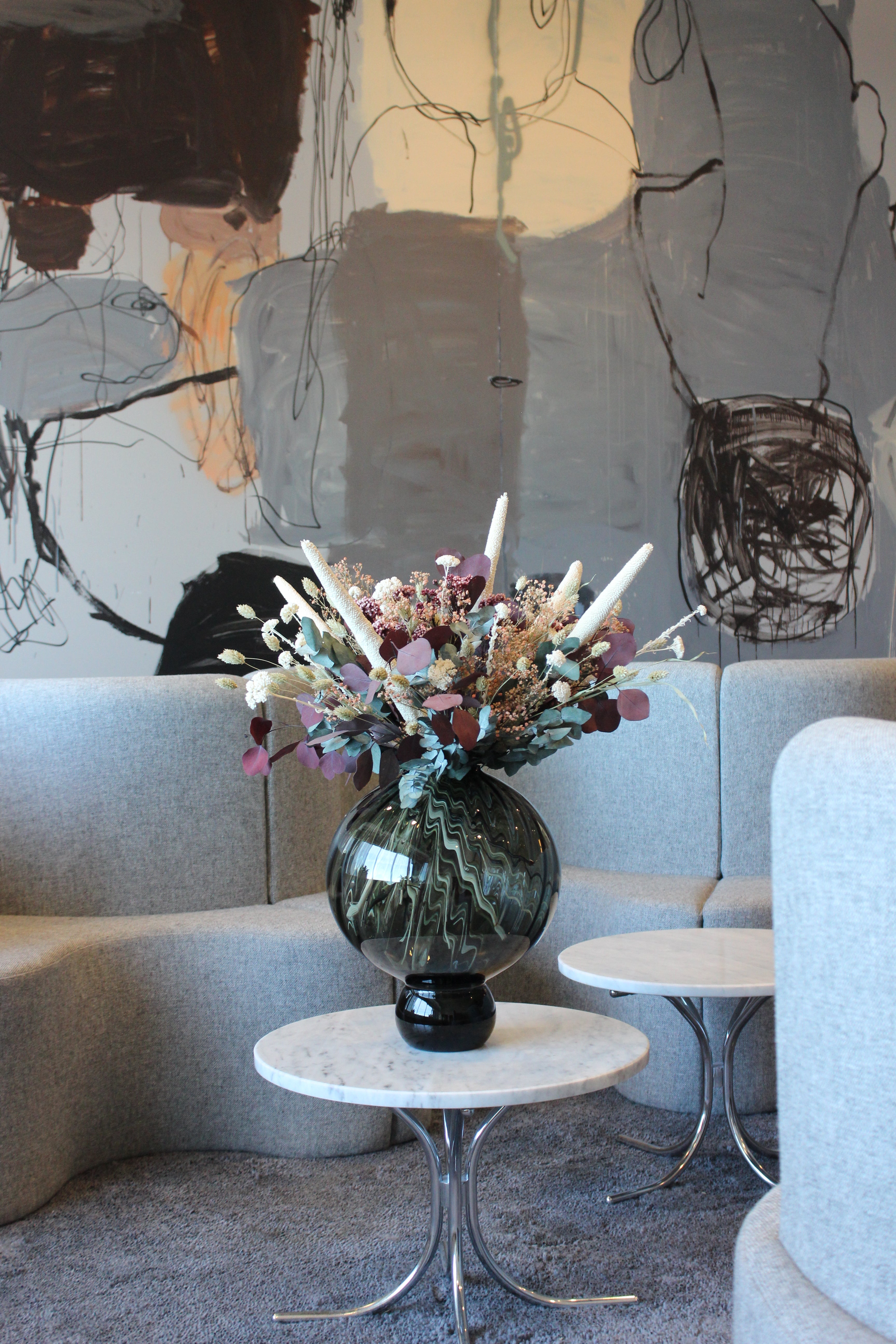 På et lille sidebord i et excentrisk indrettet rum står en stor, rund blomster-vase med en meget fyldig blomsterbuket med hvide accenter og forskellige slags eukalyptus.