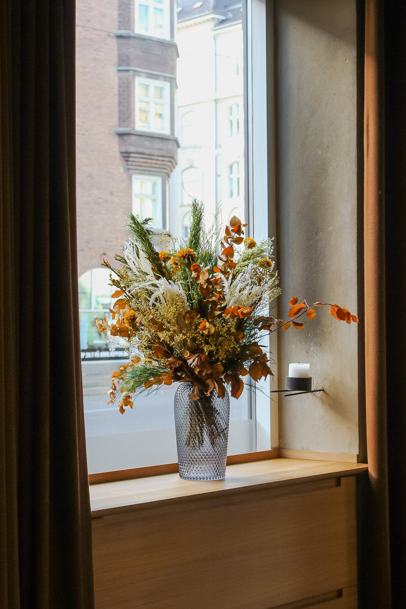 En buket i varme orange nuancer med langtidsholdbare blomster står i en glasvase i vindueskarmen.